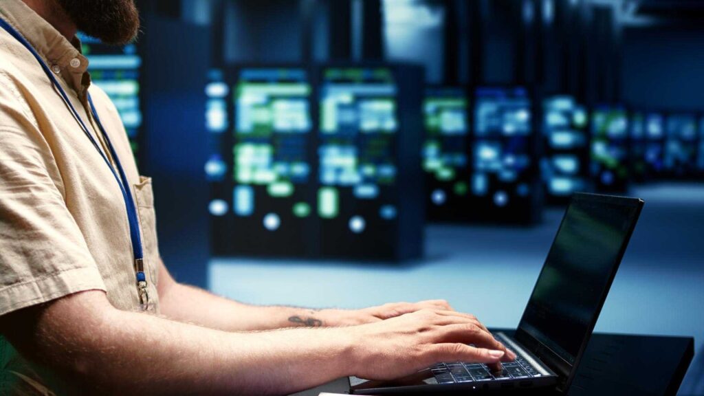 A person is working on a laptop in a dimly lit server room, with rows of illuminated server racks in the background.