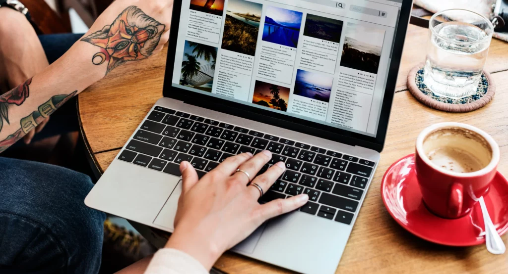 A person is using a laptop displaying a photo gallery, with a visible tattoo on one arm and a red cup on the wooden table, likely pondering the WordPress website cost for showcasing their art online. Wordpress website costs