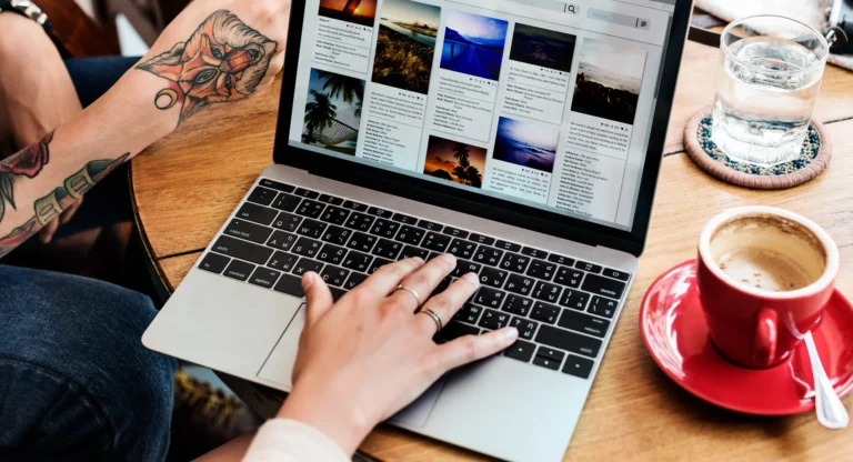 A person is using a laptop displaying a photo gallery, with a visible tattoo on one arm and a red cup on the wooden table, likely pondering the WordPress website cost for showcasing their art online. Wordpress website costs