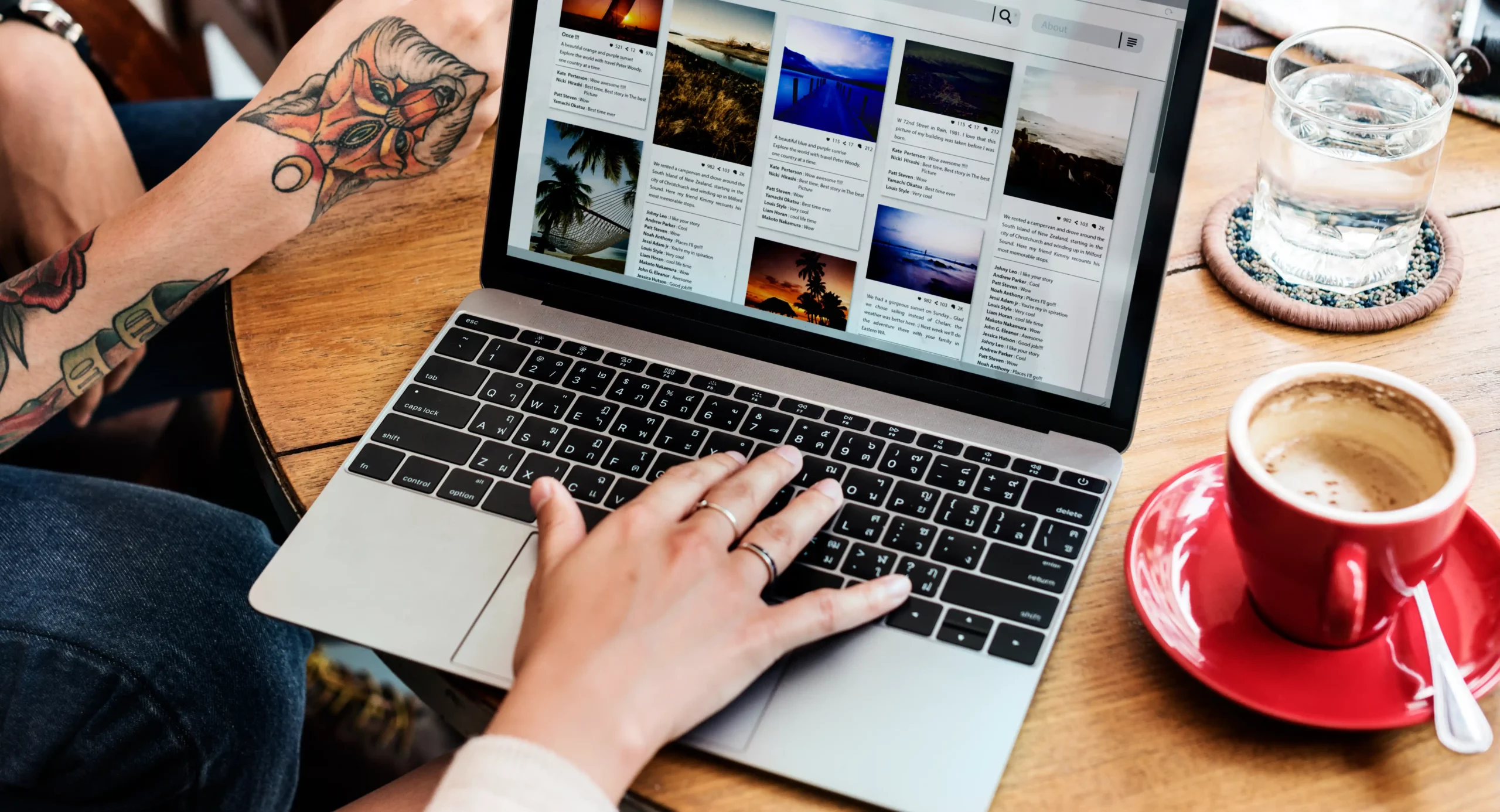 A person is using a laptop displaying a photo gallery, with a visible tattoo on one arm and a red cup on the wooden table, likely pondering the WordPress website cost for showcasing their art online. Wordpress website costs