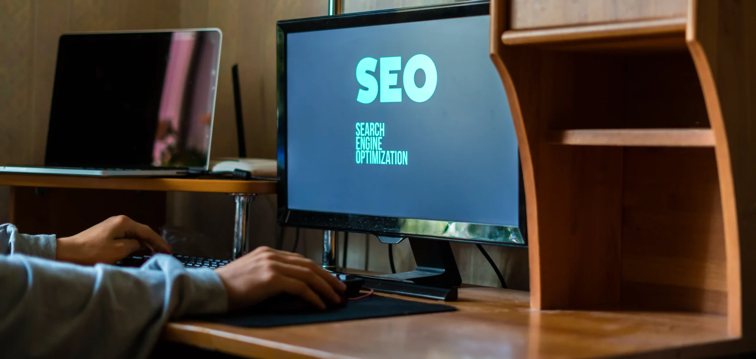 A person uses a desktop computer displaying the text "SEO Search Engine Optimization" on the screen